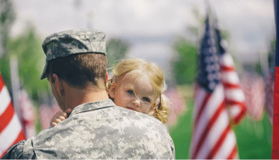 Soldier in uniform holding a young child with American flags in the background, symbolizing patriotism and family support.