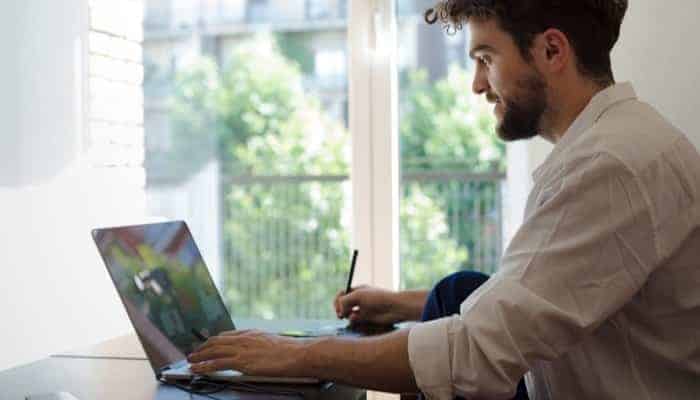 A man studying computer programming at home using a laptop and digital pen for online learning in a bright, modern space.