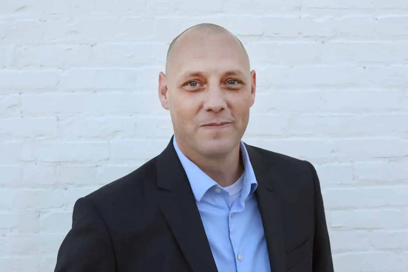Bald man in business suit smiling against a white brick wall, representing a professional figure in IT training and career development.