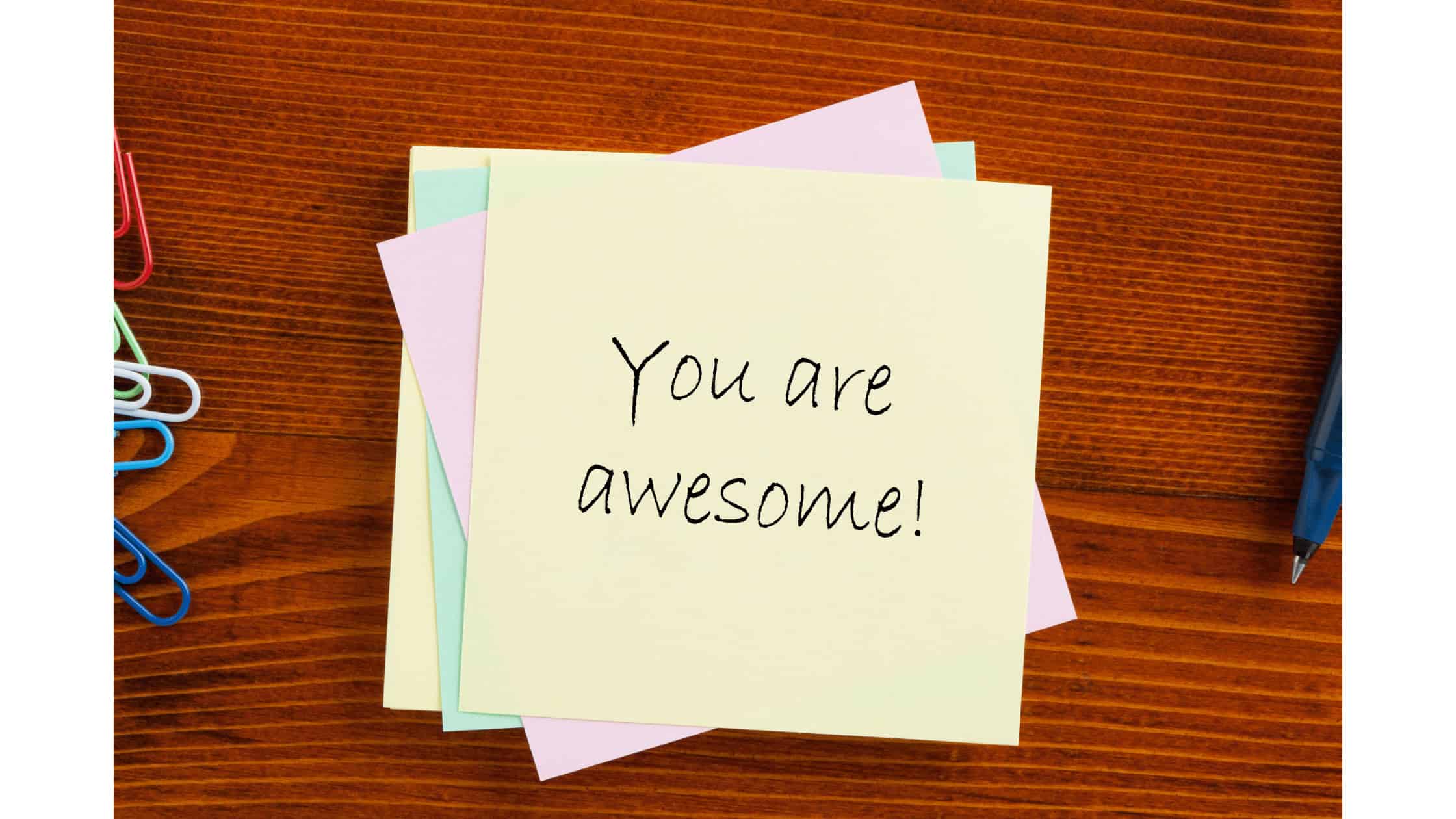 Motivational message on sticky notes with colorful paper clips, pen, and wooden desk - Inspiring workplace, positive attitude, motivational note about being awesome, professional environment, encouragement, productivity.