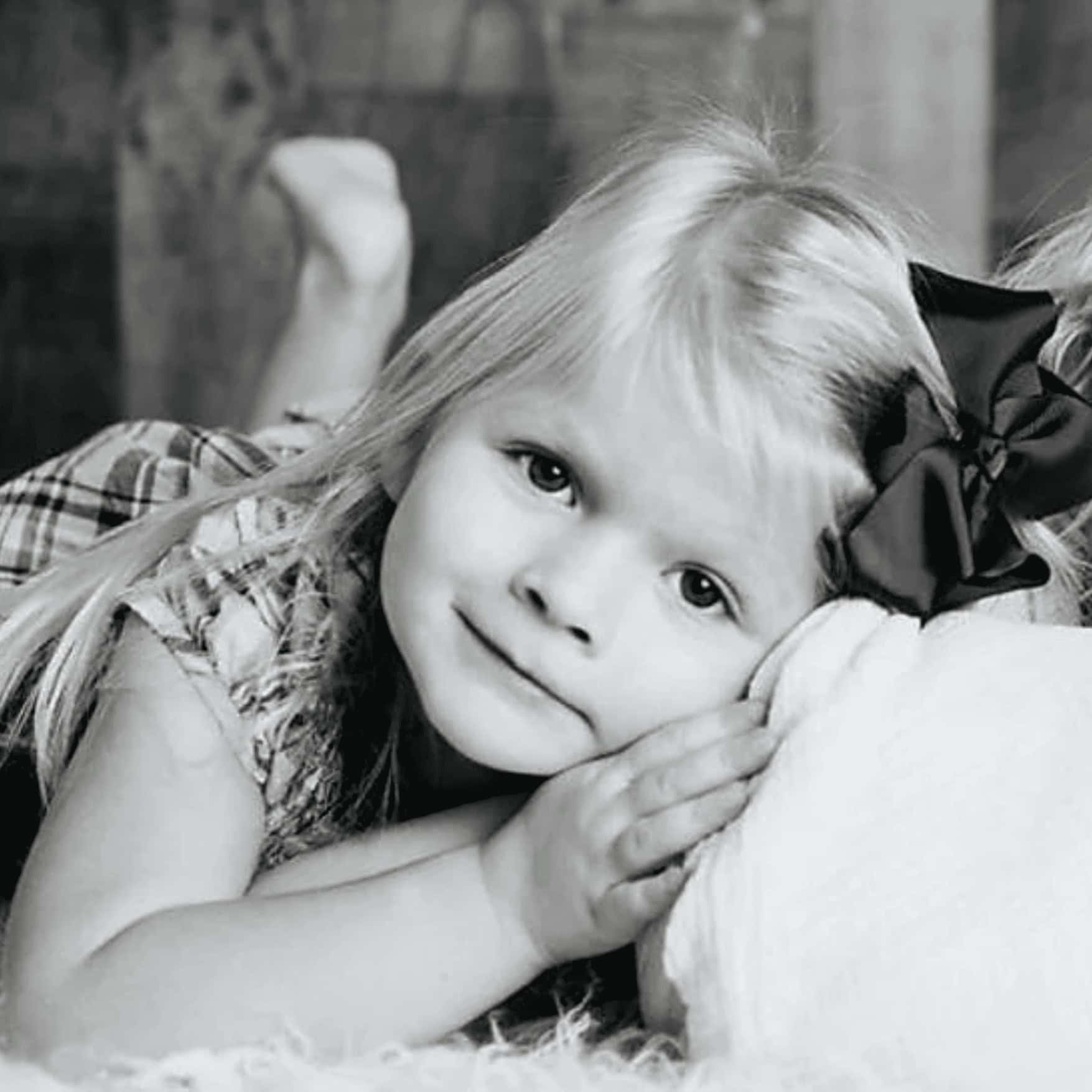 Girl lying on her side with a bow in her hair, resting her head on her hands, black and white photo showcasing innocence and childhood moments.