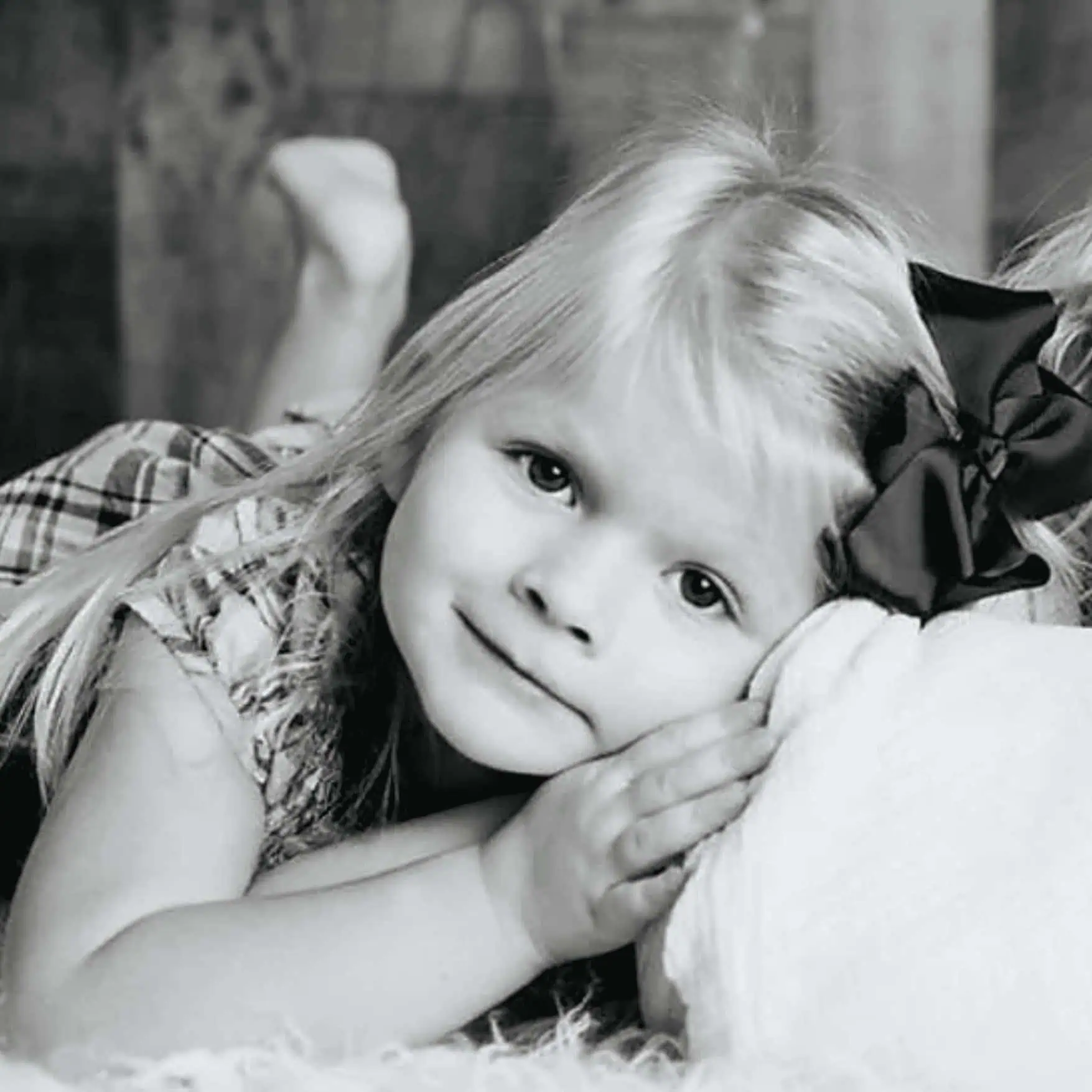Girl lying on her side with a bow in her hair, resting her head on her hands, black and white photo showcasing innocence and childhood moments.